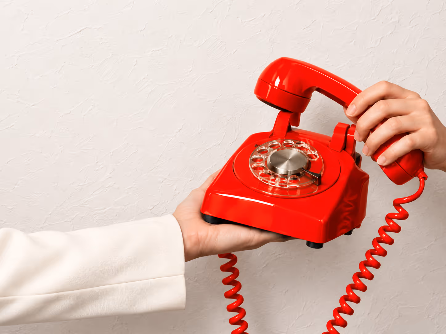 Hands holding a red rotary telephone against a light textured wall